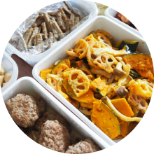 かぼちゃと蓮根の炒め物やハンバーグなどの惣菜が、タッパーに詰められて並んでいる写真